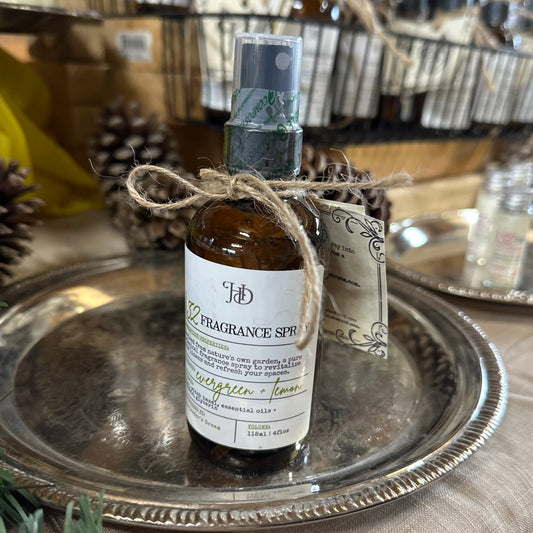A glass bottle of fragrance spray with a label that reads 'evergreen and lemon', tied with twine. The bottle is placed on a silver tray with other decorative items in the background.
