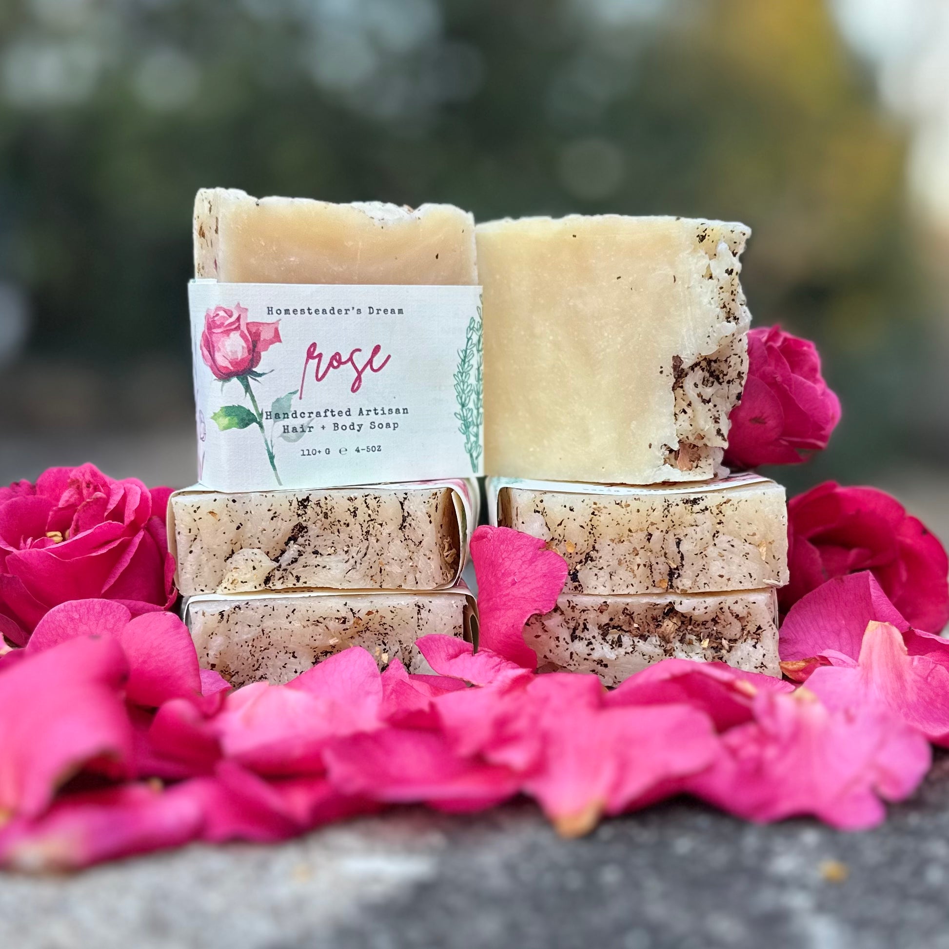 Stack of soap bars with a 'Rose' label on a bed of pink flowers outdoors.