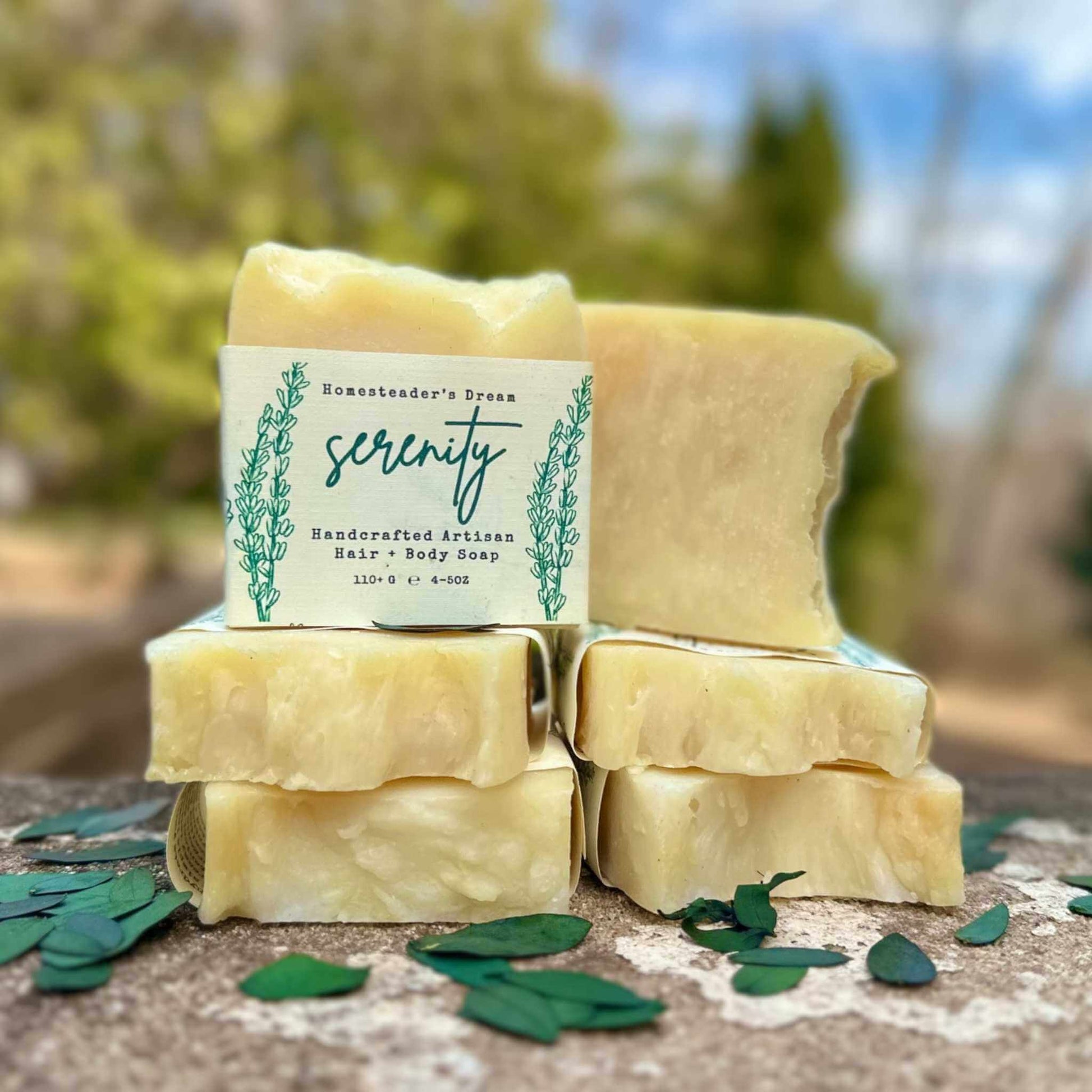 Stack of handmade soap bars with a 'serenity' label on a natural surface with greenery in the background.