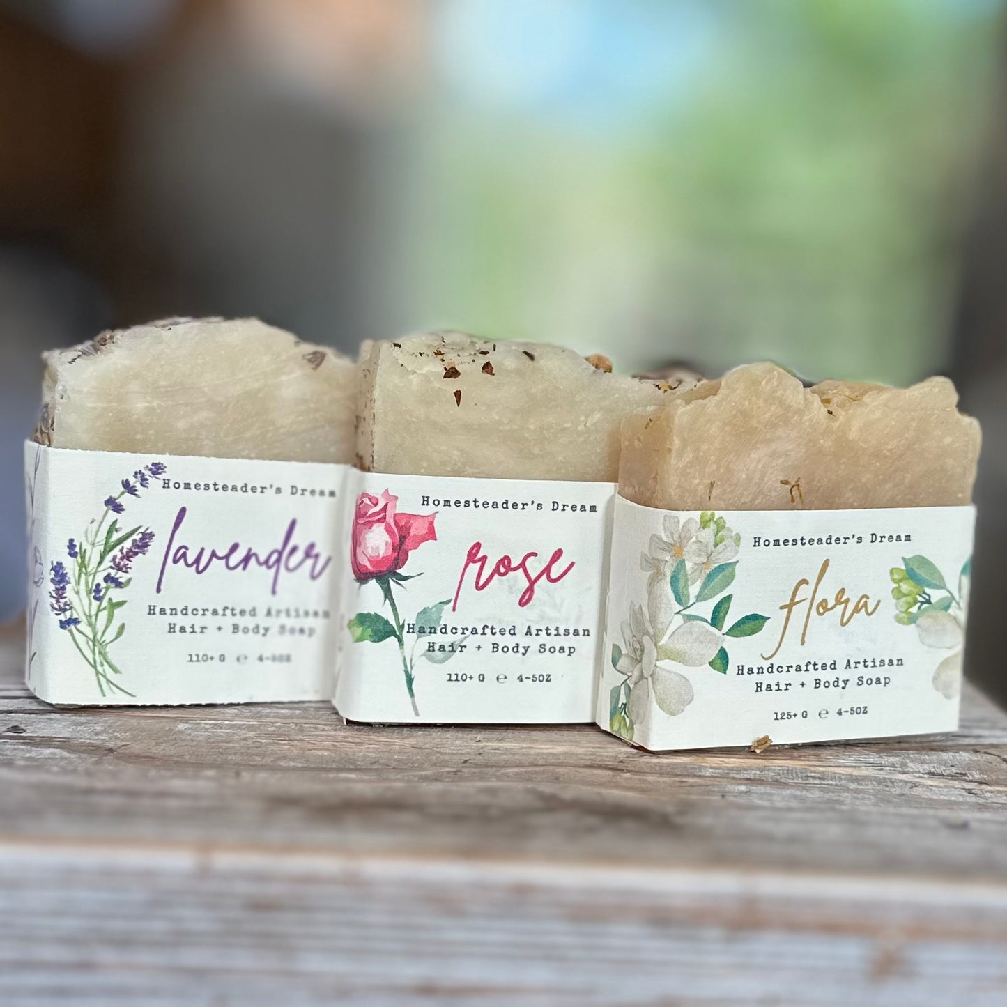 Three bars of soap with labels for lavender, rose, and flora on a wooden surface.