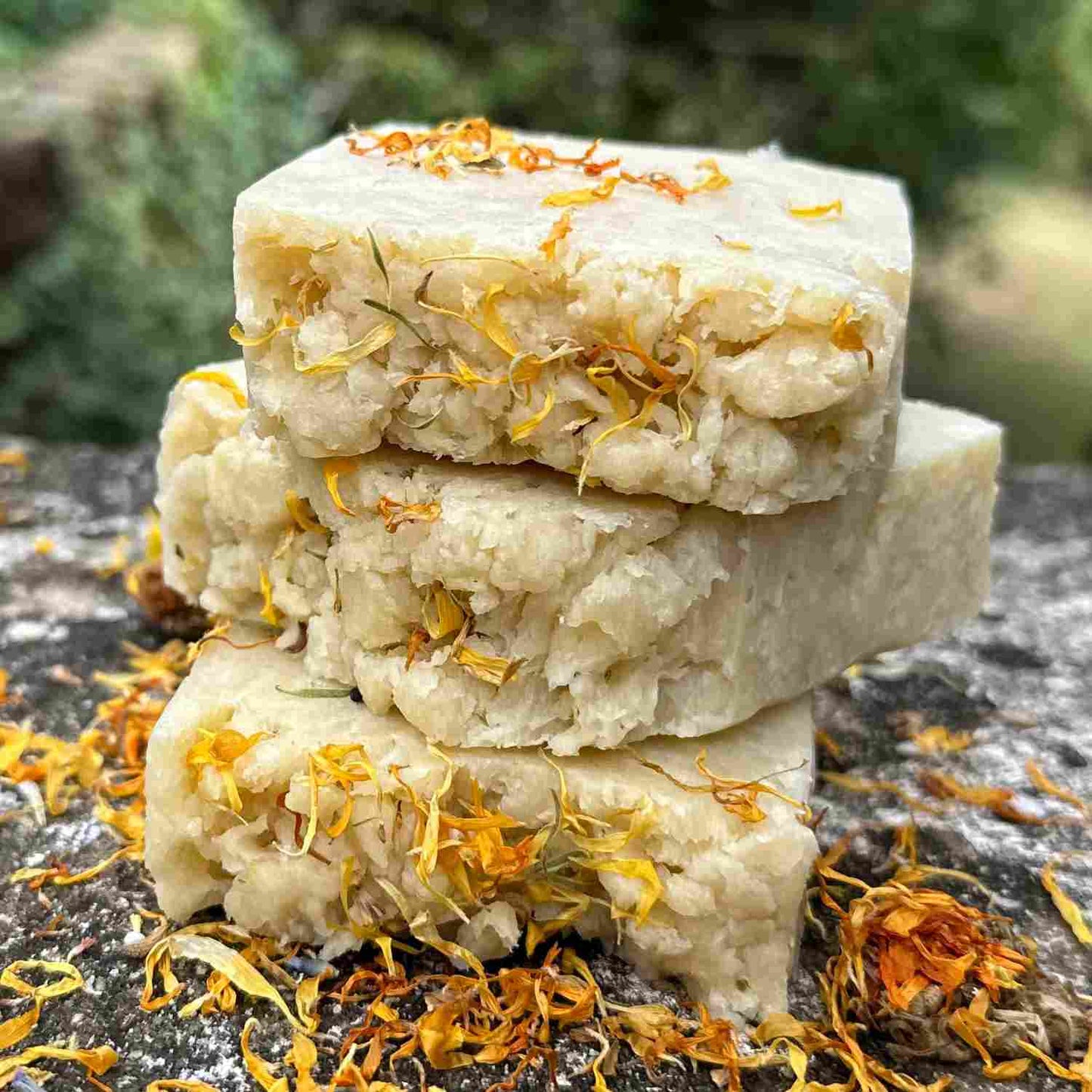Stack of three artisan-crafted soaps with yellow calendula flower petals on top, placed on a natural stone surface.