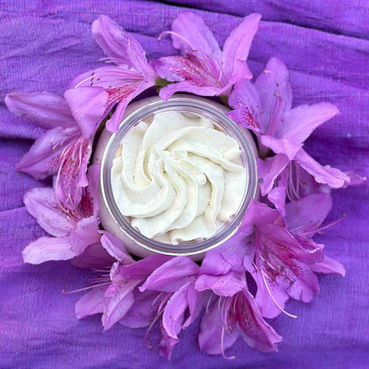 A container of Luxurious Lavender Body Butter surrounded by purple flowers, with a purple fabric in the background.