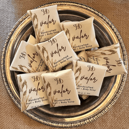 Several individually wrapped bars of Valor Handcrafted Hair & Body Soap arranged on a decorative round metal tray over a burlap-like surface.
