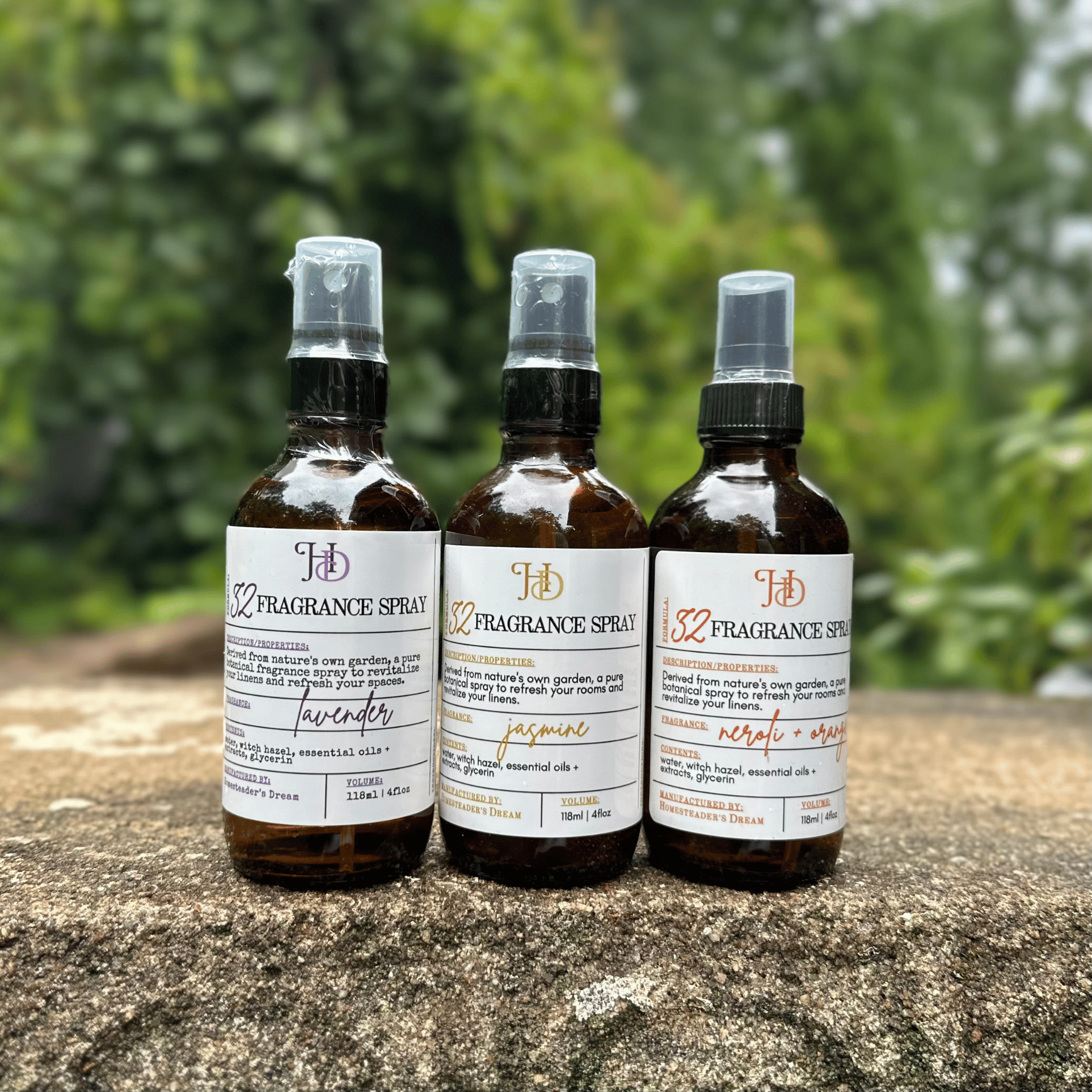 Three amber glass fragrance spray bottles with apothecary labels are lined up on a stone surface outdoors, with a blurred green garden background.