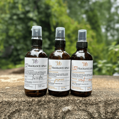 Three amber glass fragrance spray bottles with apothecary labels are lined up on a stone surface outdoors, with a blurred green garden background.
