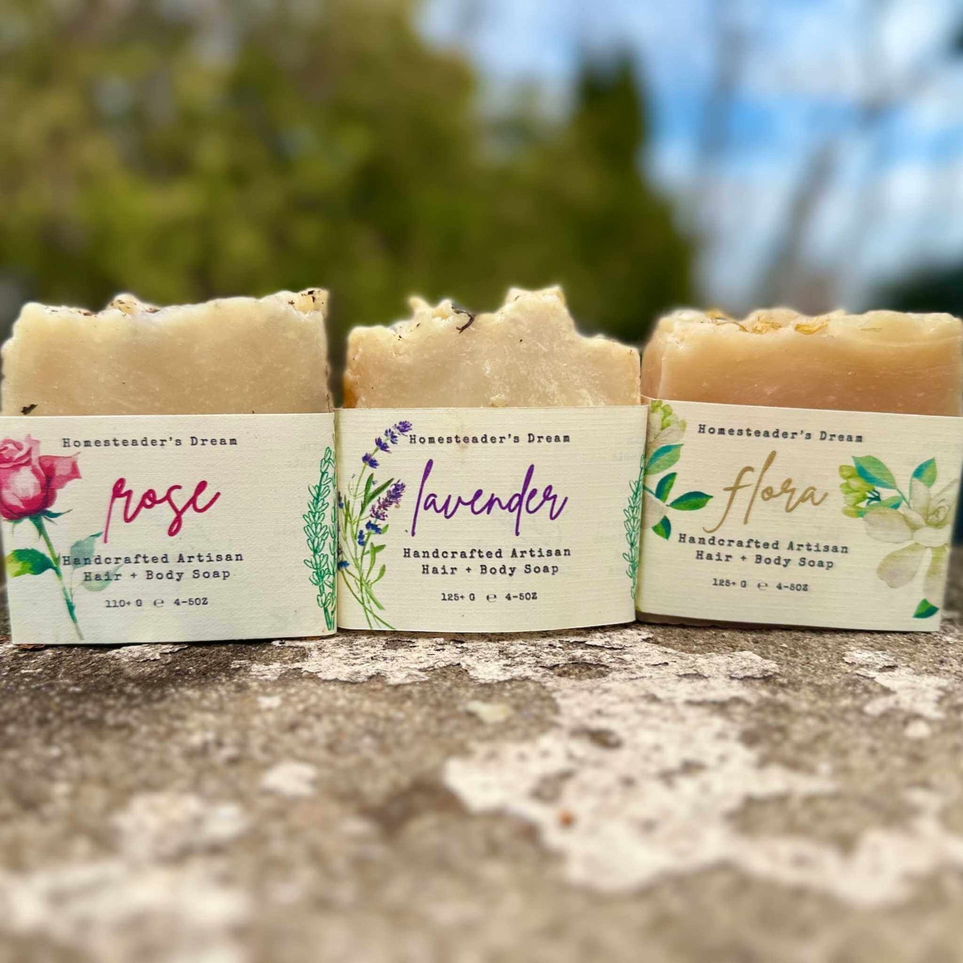 Three bars of soap with labels for 'Rose', 'Lavender', and 'Flora' on a stone surface.