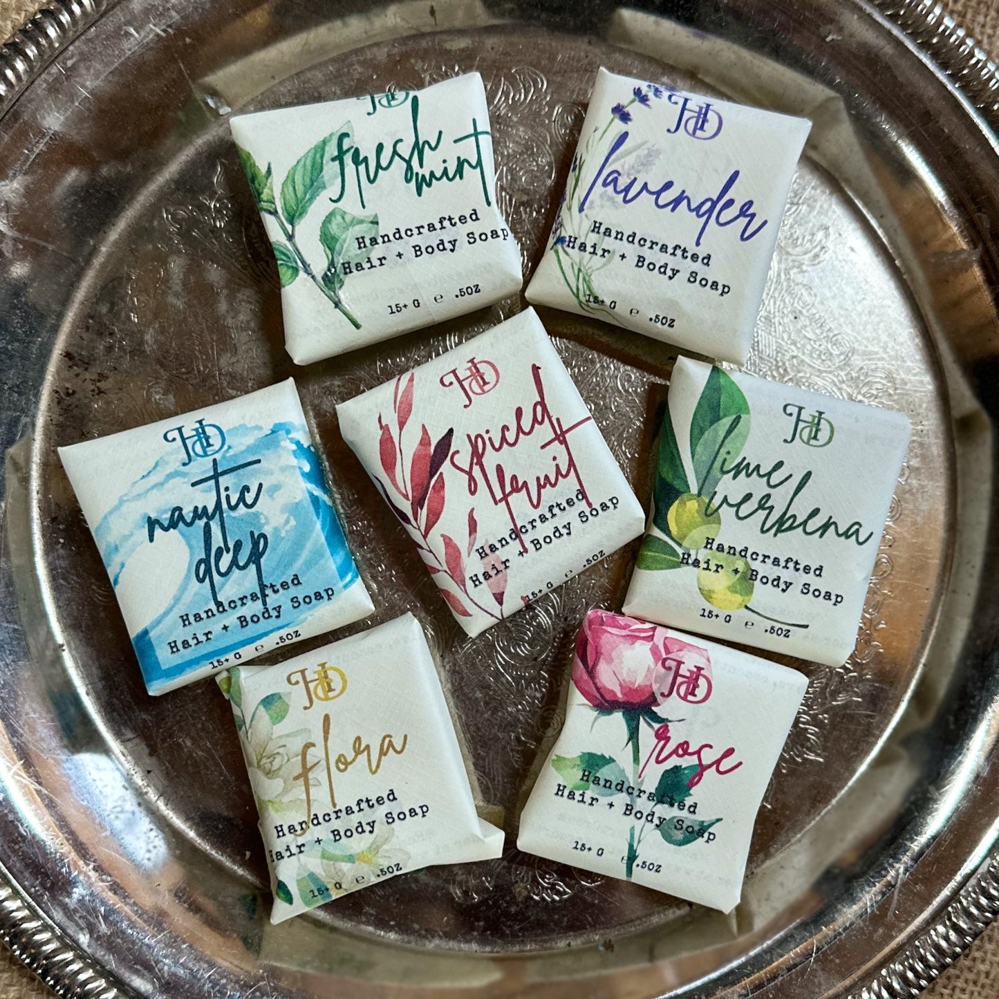 Assorted handcrafted hair and body guest soaps with designer labels on a silver tray.