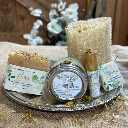 Collection of natural beauty products including soaps, body butter, a perfume and a loofah on a textured surface with a blurred floral background.