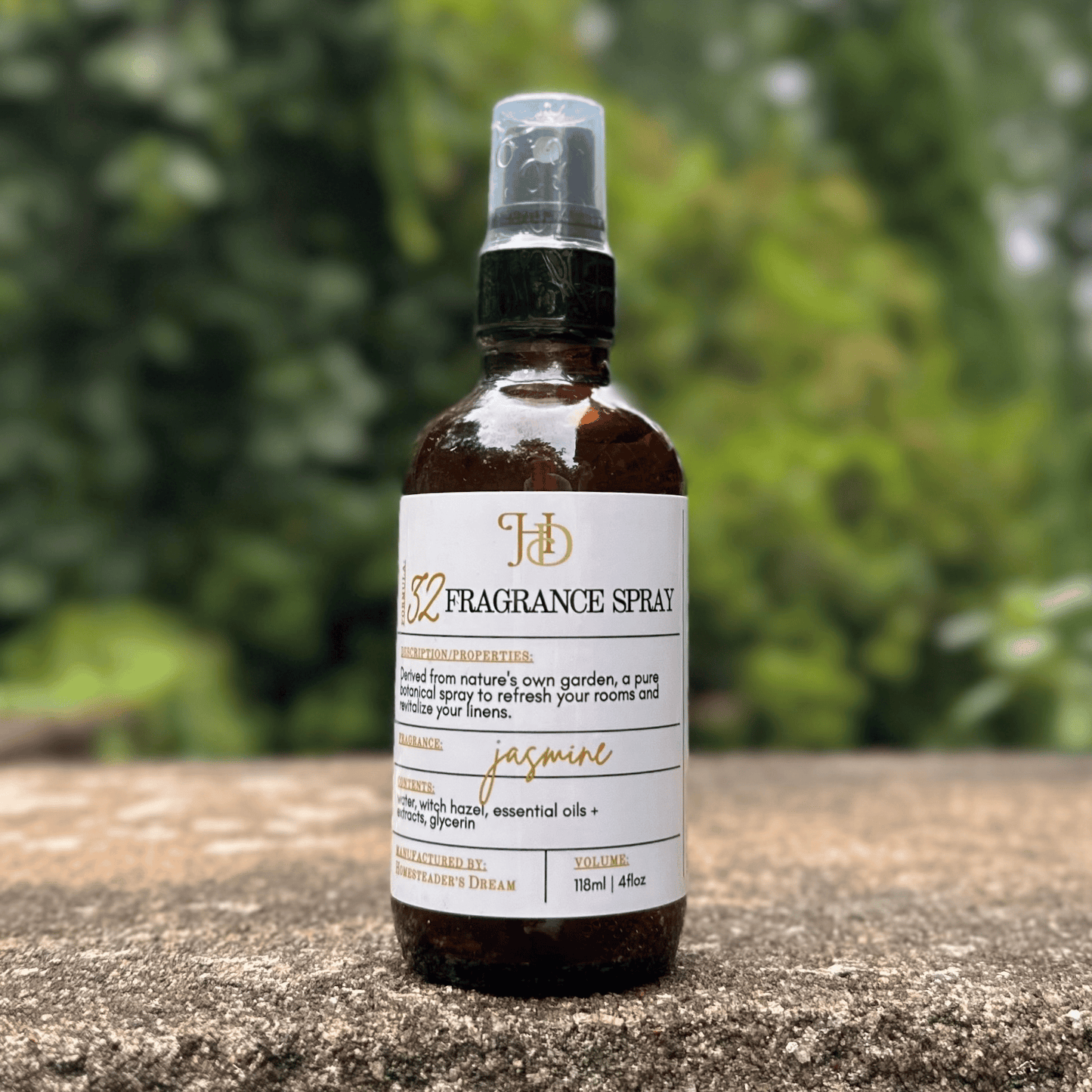 A 4 oz amber glass fragrance spray bottle with an apothecary label reading 'jasmine', standing on a textured stone surface with a blurred green garden background.