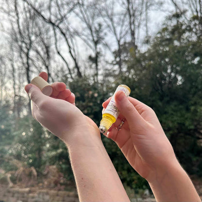 Two hands holding small bottles against a natural background with trees and greenery.