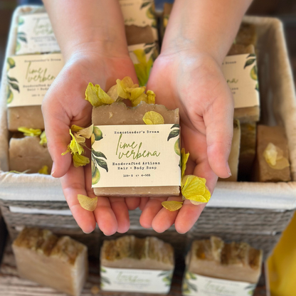 Hands holding a bar of lime verbena soap with a linen paper label, surrounded by more soap bars in the background.