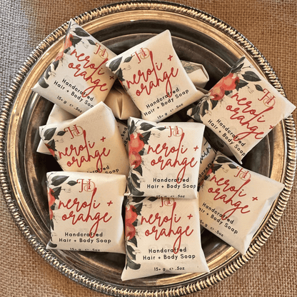 Several wrapped bars of hair and body soap arranged on a round decorative metal tray over a burlap surface. The soap wrappers are cream with orange script reading 'neroli + orange' and botanical illustrations.