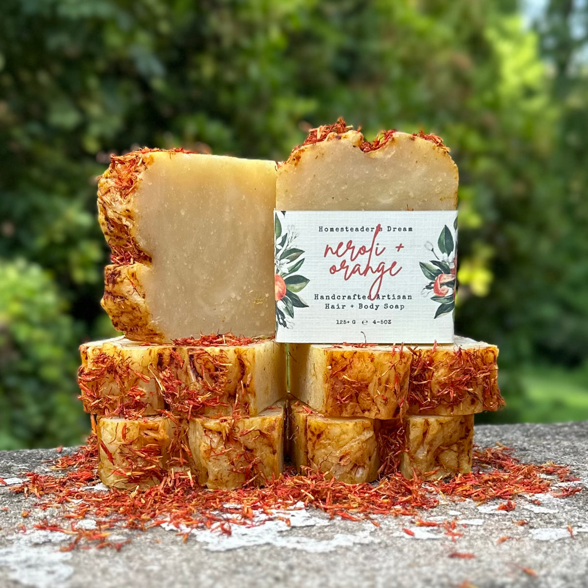Six bars of handmade hair and body soap  in the 'neroli + orange' scent, stacked on a stone surface with a blurred green garden background. The soap bars are a warm cream-beige color and are sprinkled with dried orange petals.