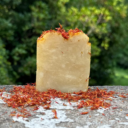 A rectangular bar of handmade soap with a pale beige body and orange dried flower petals on top, standing on a stone surface with more petals scattered around it against a blurred green outdoor background.