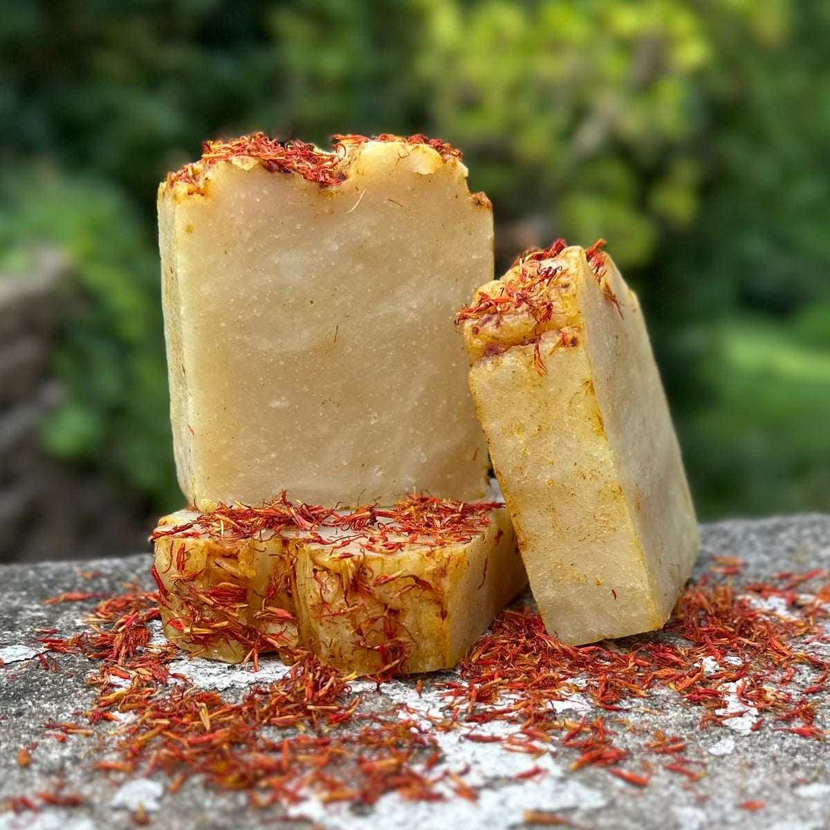 Three handmade bars of soap with orange petals on top, displayed on a stone surface with a blurred green garden background.