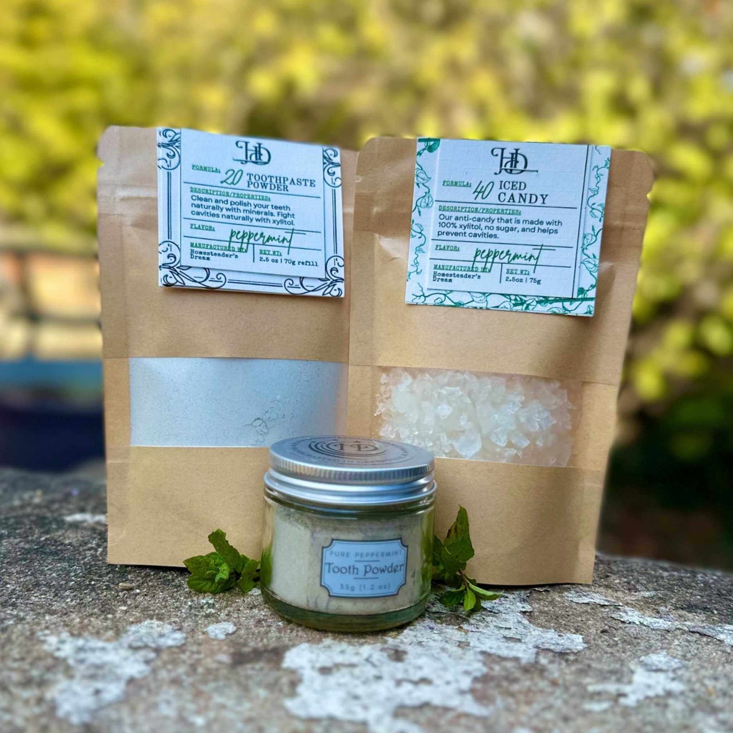 Two brown paper bags with labels and a small jar of tooth powder on a stone surface with a blurred green background.