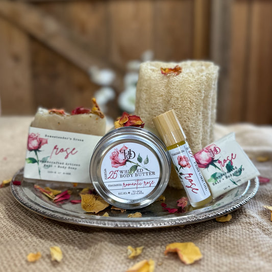 Collection of real rose oil products including a body butter, perfume, and soaps on a silver tray on a burlap cloth sprinkled with rose petals with a wooden background.