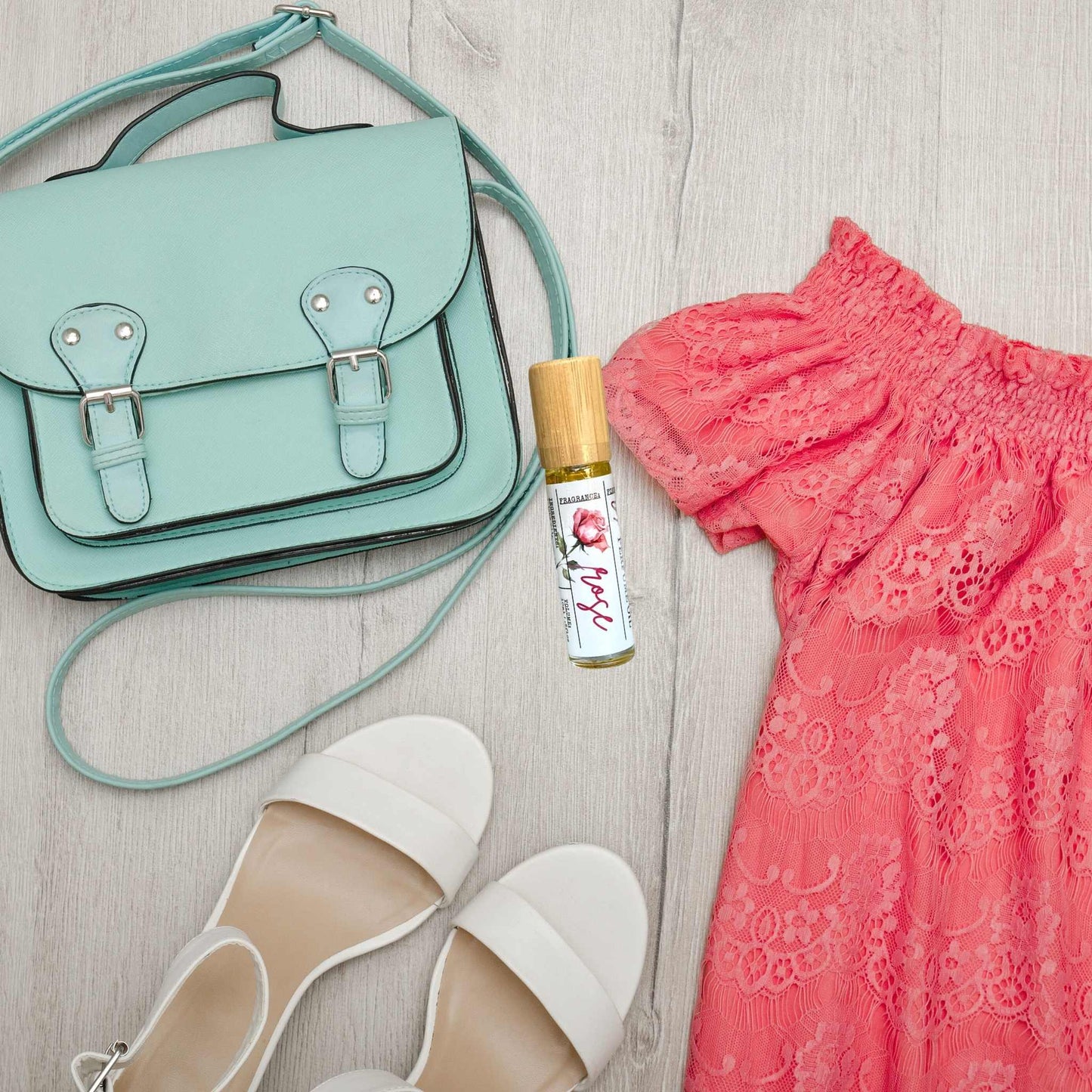 Mint green handbag, white sandals, pink lace dress, and a bottle of perfume on a light wooden surface.