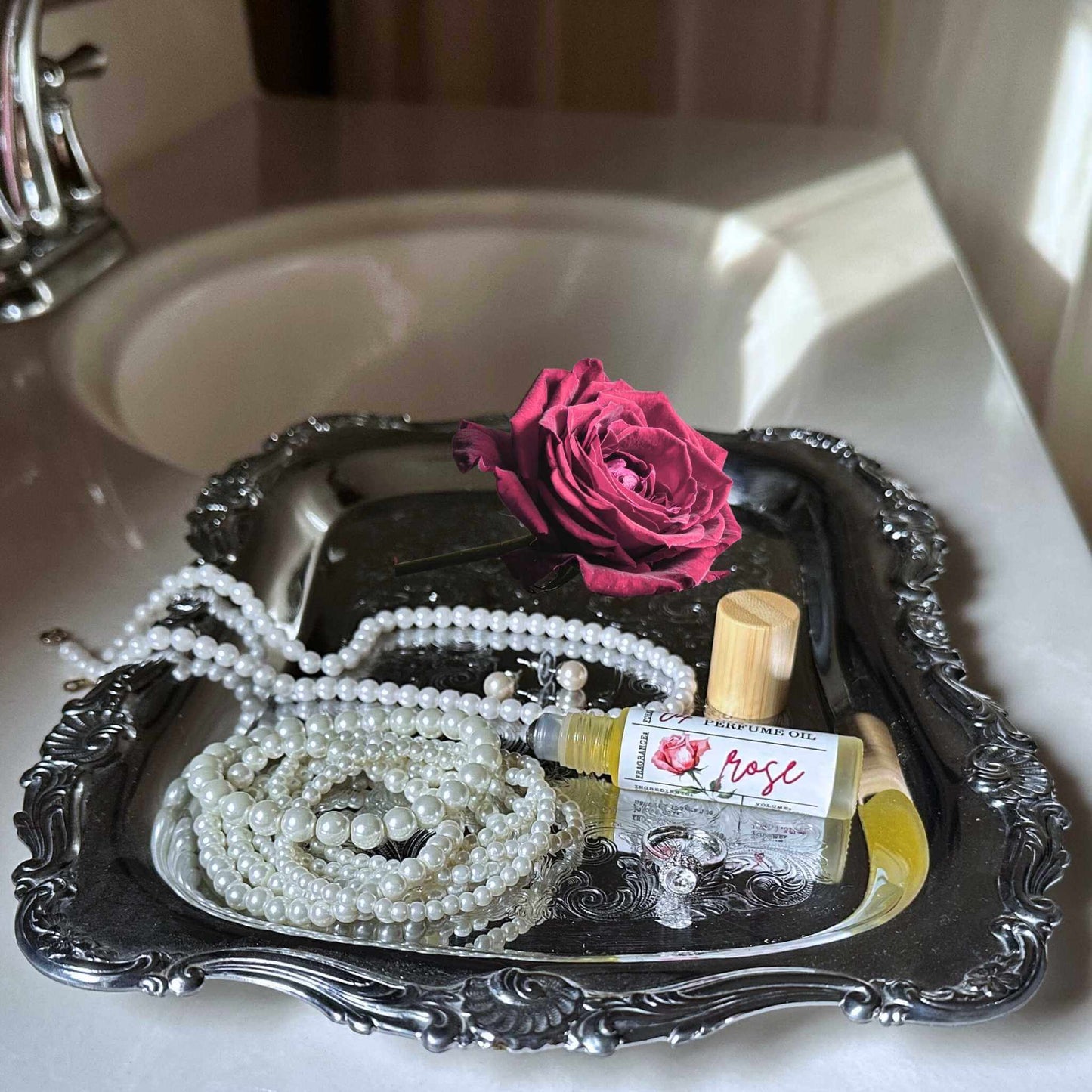 Tray with pearls, a bottle of rose perfume, and a pink rose on a sink.