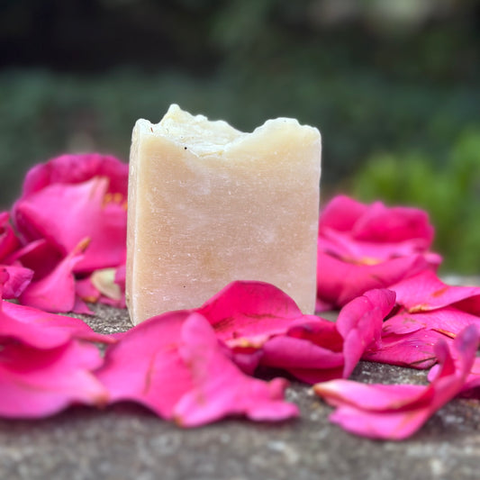 A bar of soap resting on a bed of pink rose petals, with a blurred green background.