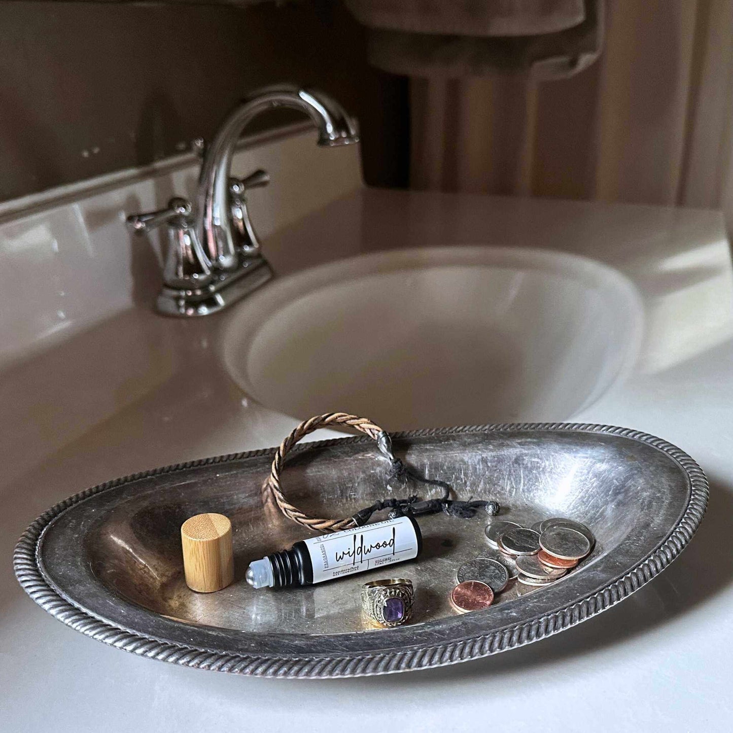 Silver tray with personal items including a roll-on bottle, wooden lid, and coins on a bathroom sink.